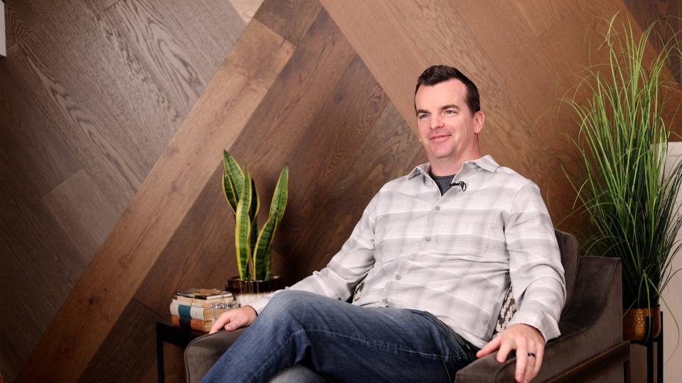 A man sits in a chair relaxed wearing a button up plaid flannel.
