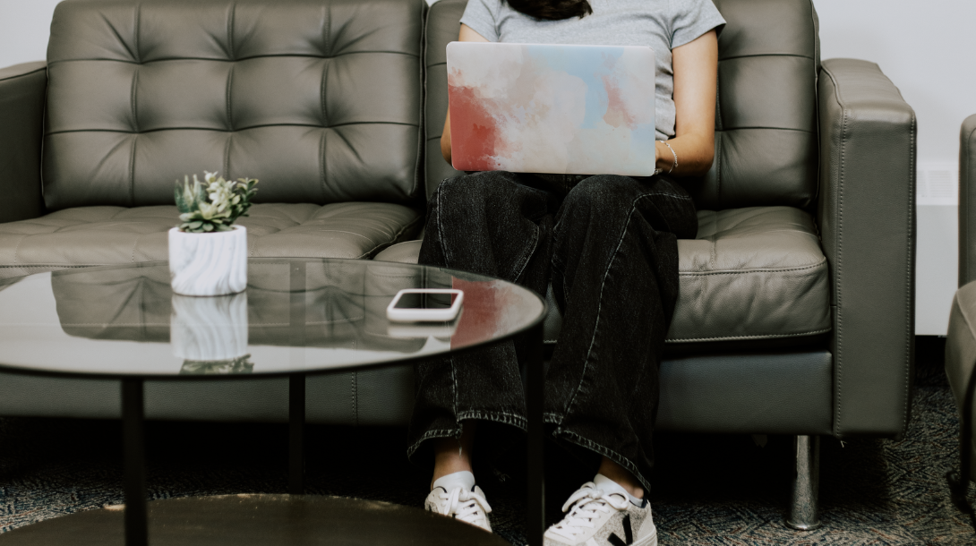 A person sitting on a pleather couch with laptop in lap and phone on a coffee table, looking up volunteering opportunities.
